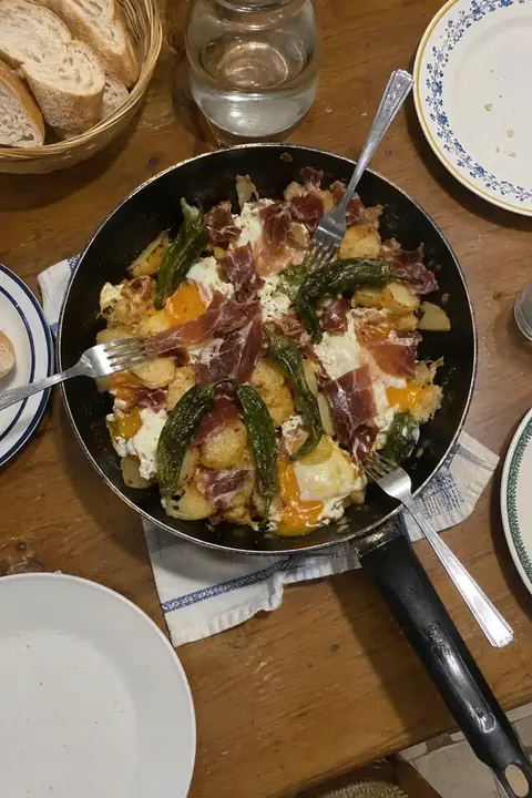 Sartén de huevos rotos con jamón y pimientos sobre la mesa del comedor, con tres tenedores ya pinchados